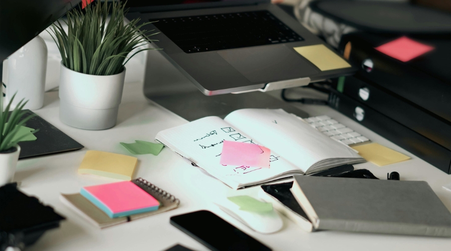 Neredan radni sto pretrpan sveskama, papirićima, telefonima, fasciklama i laptopom podignutim na stalku, okružen sobnim biljkama.