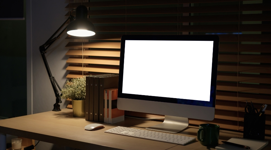 Uredan radni sto sa velikim monitorom koji ima bel ekran, stolnom lampom, knjigama, tastaturom i šoljom, u prostoru sa zatvorenim žaluzinama.