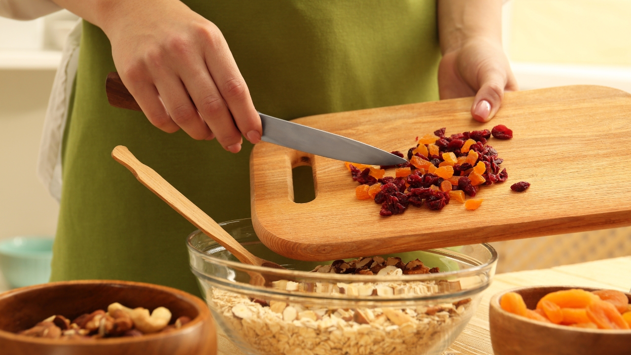 Osoba seče suvo voće i dodaje ga u činiju sa ovsenim pahuljicama i orašastim plodovima