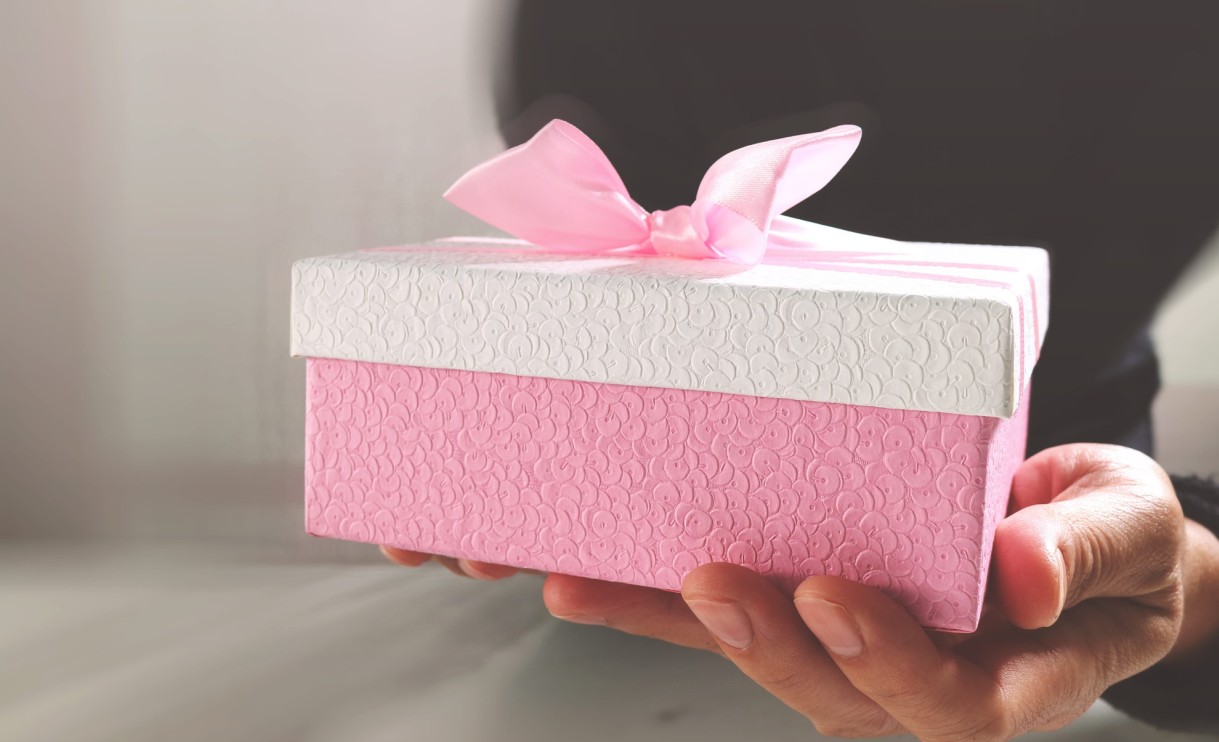 Ruke drže roze-belu ukrasnu kutiju sa mašnom, pripremljenu kao poklon za ženu